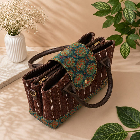A structured brown woven handbag with dark brown leather handles and trim, featuring a green and orange patterned fabric flap and matching patterned base, styled on a beige surface with decorative plants and a vase.