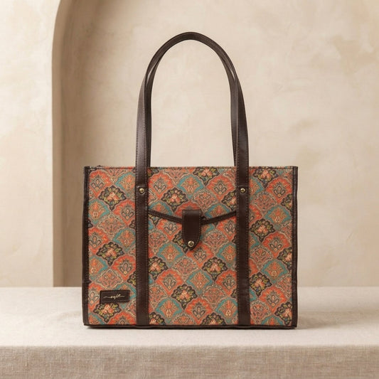 A structured tote bag with a geometric patterned body in rust orange, teal, and black, accented by dark brown leather handles and trim, displayed on a neutral beige surface.