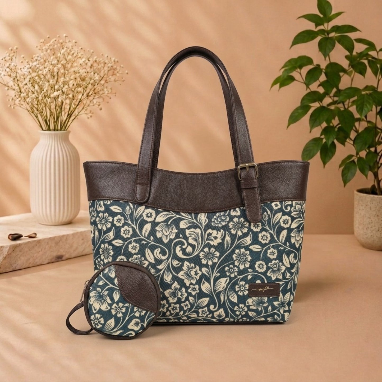 A tote bag with a dark floral print and dark brown faux-leather trim, shown with a matching small pouch. The bag is styled on a beige surface with a vase of dried flowers and a potted plant in the background.