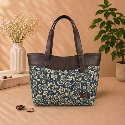 A tote bag with a navy blue and cream floral patterned body and dark brown faux-leather trim and handles, displayed on a beige surface with decorative props including a vase of dried flowers and a potted plant.