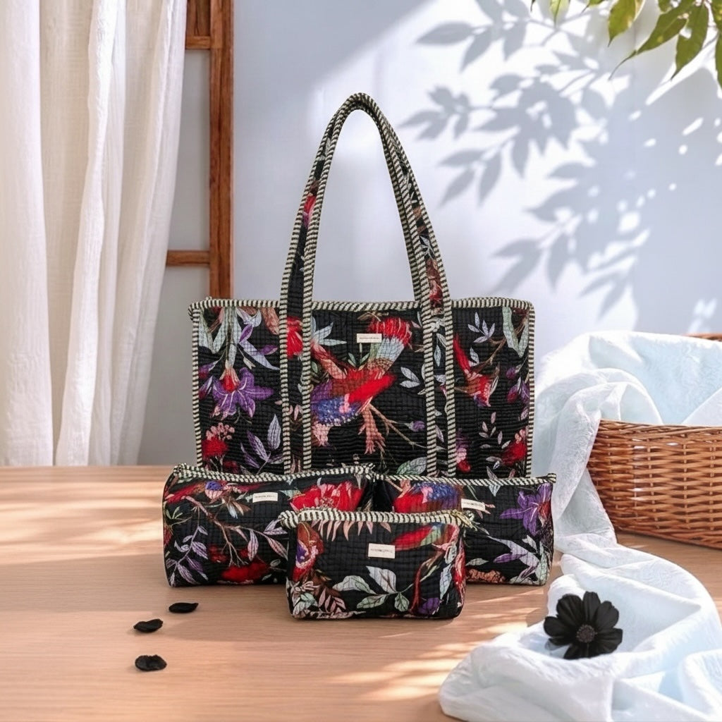 A matching tote bag and two zip pouches with a black background and a colorful floral print featuring red birds and purple flowers, styled on a wooden table with a basket and white fabric in a sunlit room.