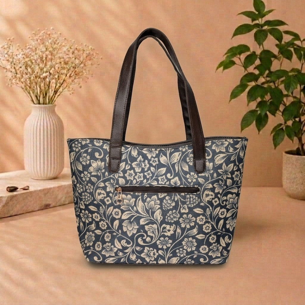 A navy blue tote bag with a cream floral pattern and black handles, displayed on a beige surface with decorative plants and a vase in the background.