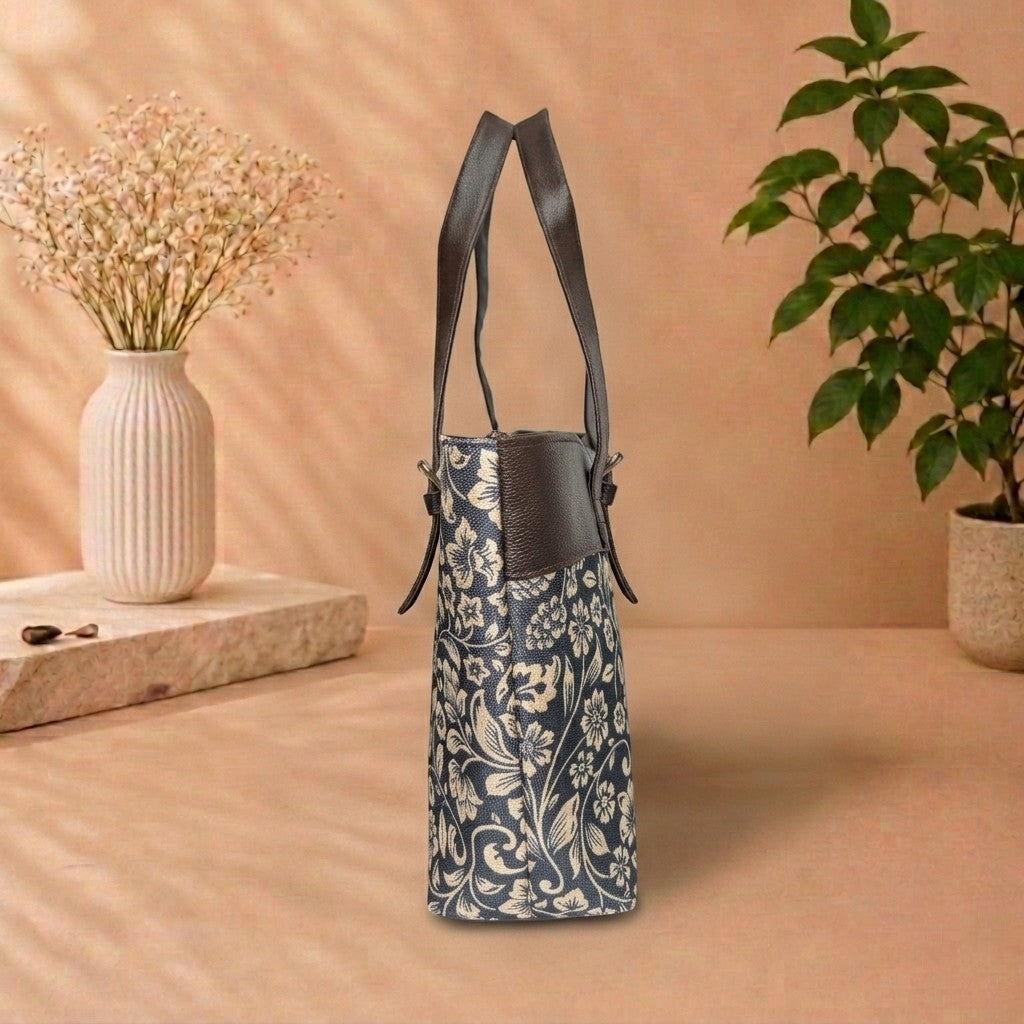 A navy blue and beige floral patterned handbag with dark brown leather shoulder straps, standing on a beige surface with a vase of dried flowers and a potted plant in the background.