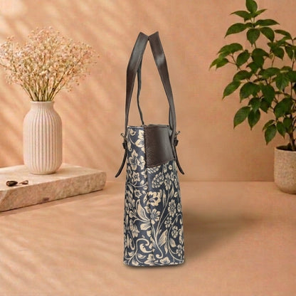A navy blue and beige floral patterned handbag with dark brown leather shoulder straps, standing on a beige surface with a vase of dried flowers and a potted plant in the background.