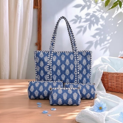A matching blue and white ikat patterned bag set on a wooden table, featuring a tote bag, a medium zippered pouch, and a small zippered pouch. The set is styled with white draped fabric, a wicker basket, and soft natural shadows.