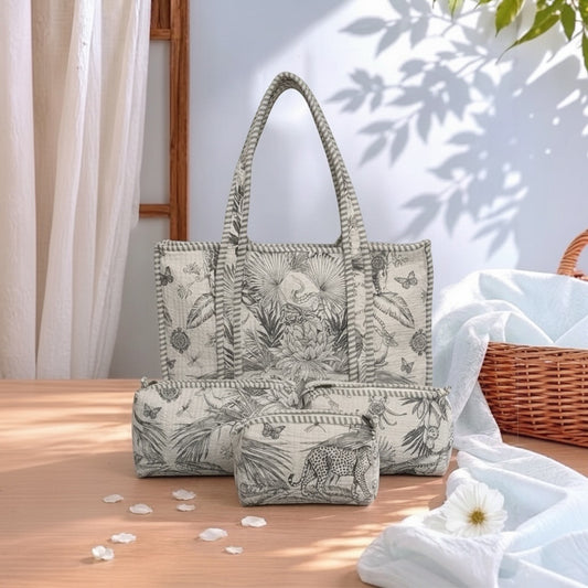A coordinated tote bag, matching pouch, and small zip pouch set in a tropical leaf and animal print, styled on a wooden table with draped white fabric, a wicker basket, and soft natural shadows.