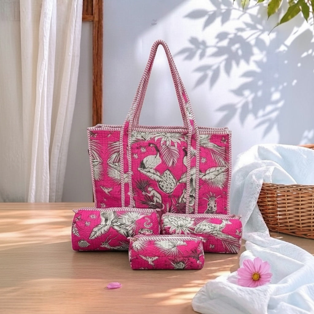 A coordinated set of bright pink tropical-print fabric accessories, including a tote bag, a rolled mat or blanket, and two small pouches, arranged on a wooden table with draped white fabric, a wicker basket, and a pink flower nearby.