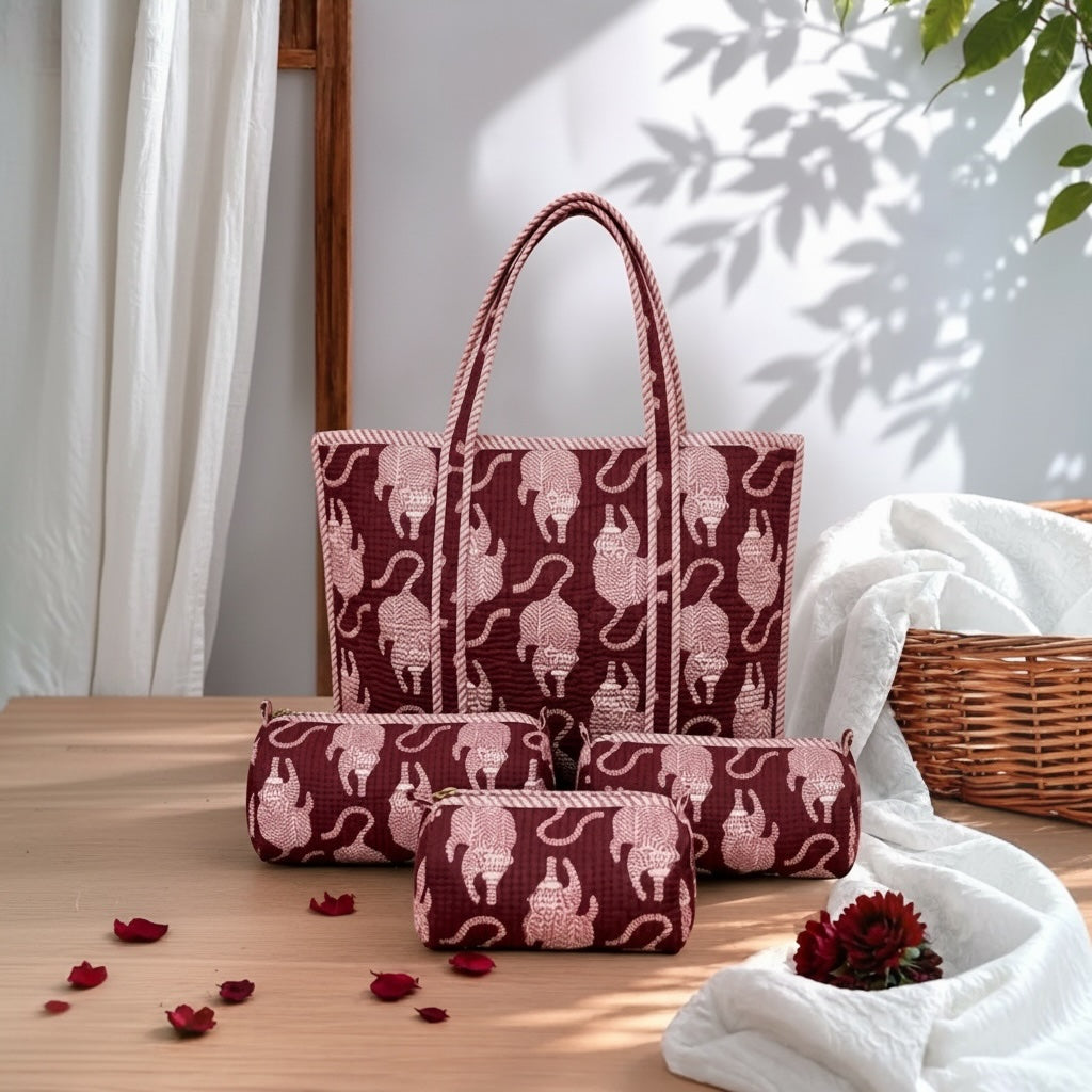 A coordinated set of fabric tote bag and small pouches in a deep burgundy color with a light pink cat pattern, arranged on a wooden table with white fabric, curtain, and wicker basket in a styled indoor setting.