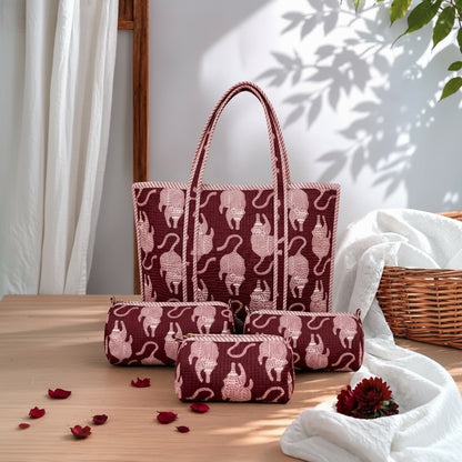 A coordinated set of fabric tote bag and small pouches in a deep burgundy color with a light pink cat pattern, arranged on a wooden table with white fabric, curtain, and wicker basket in a styled indoor setting.