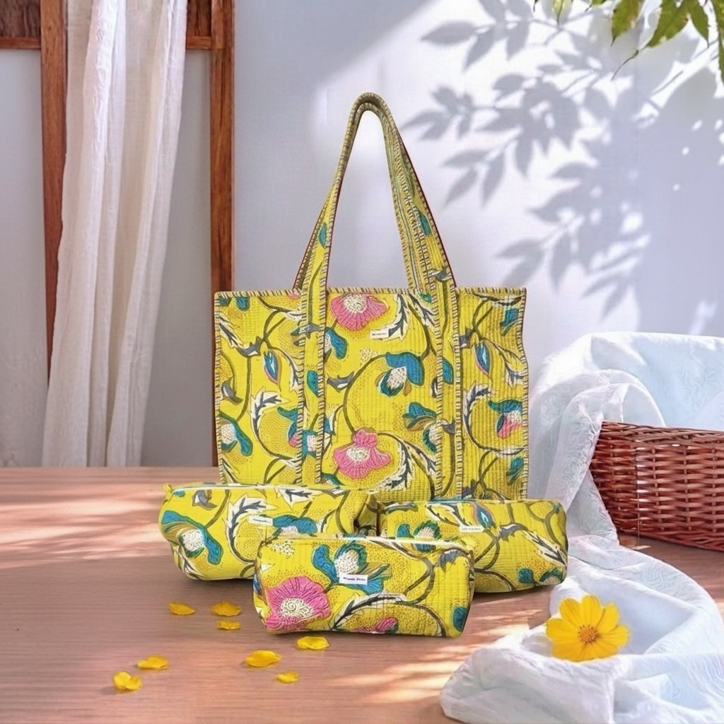 A bright yellow fabric tote bag with a floral print, shown alongside matching rolled-up pouches on a wooden table. The print features pink flowers, green leaves, and blue accents.