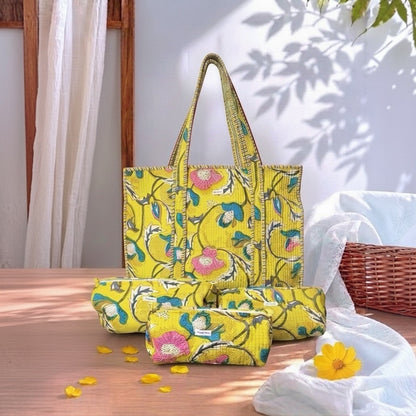A bright yellow fabric tote bag with a floral print, shown alongside matching rolled-up pouches on a wooden table. The print features pink flowers, green leaves, and blue accents.