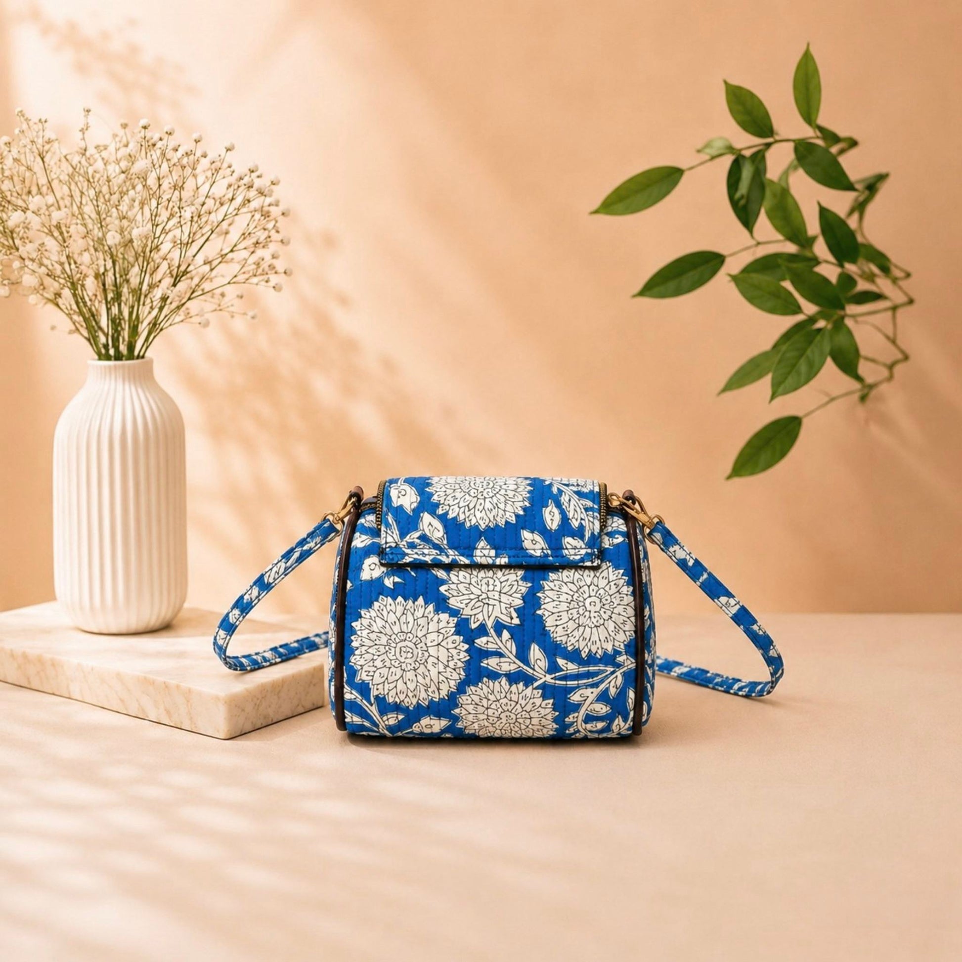 Blue floral-patterned handbag on a beige surface with a vase and plant in the background