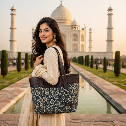 A woman in a cream-colored traditional outfit stands in front of the Taj Mahal, carrying a large navy blue tote bag with a beige floral pattern and dark brown leather trim.