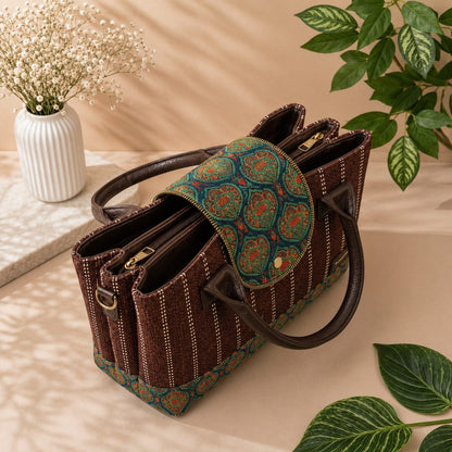 A structured brown woven handbag with dark brown leather handles and trim, featuring a green and orange patterned fabric flap and matching patterned base, styled on a beige surface with decorative plants and a vase.