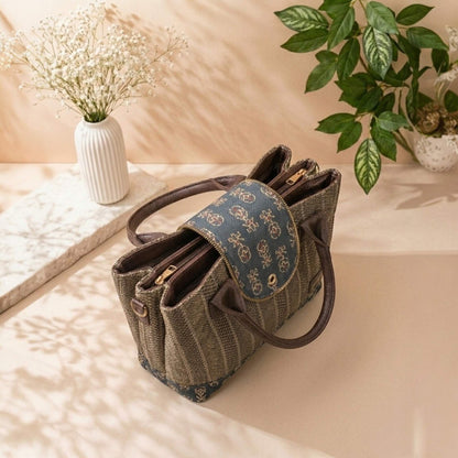 Brown textured handbag with decorative flap on a light surface with plants in the background
