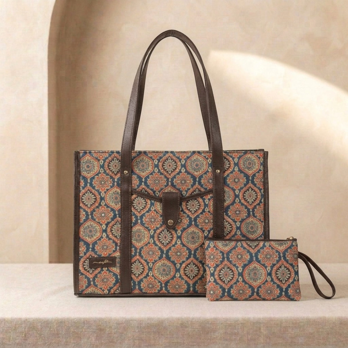 A patterned tote bag with dark brown leather handles and trim, shown alongside a matching zip pouch on a beige surface against a softly lit neutral background.