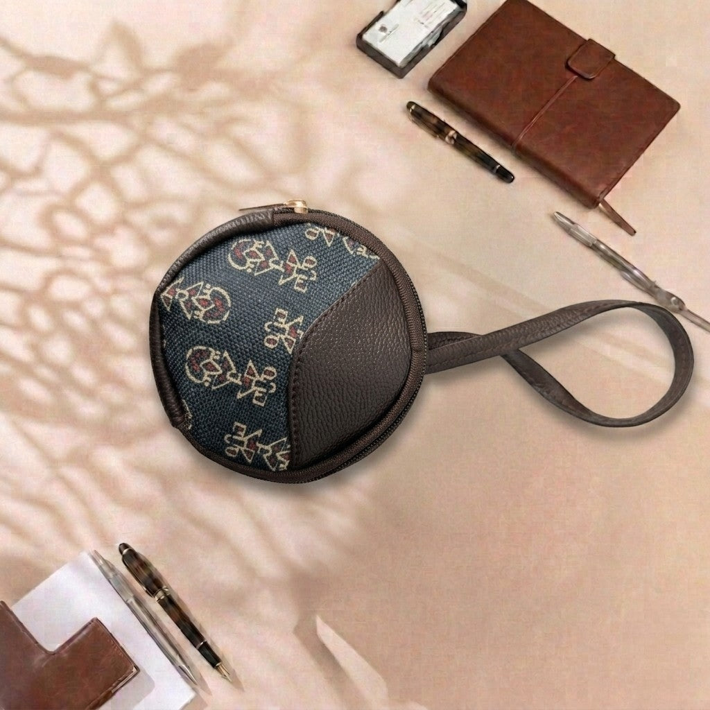 A small round zip pouch with a dark brown leather strap and trim, featuring a blue fabric front with a beige floral pattern, styled on a beige surface with a brown notebook, pen, and other accessories.