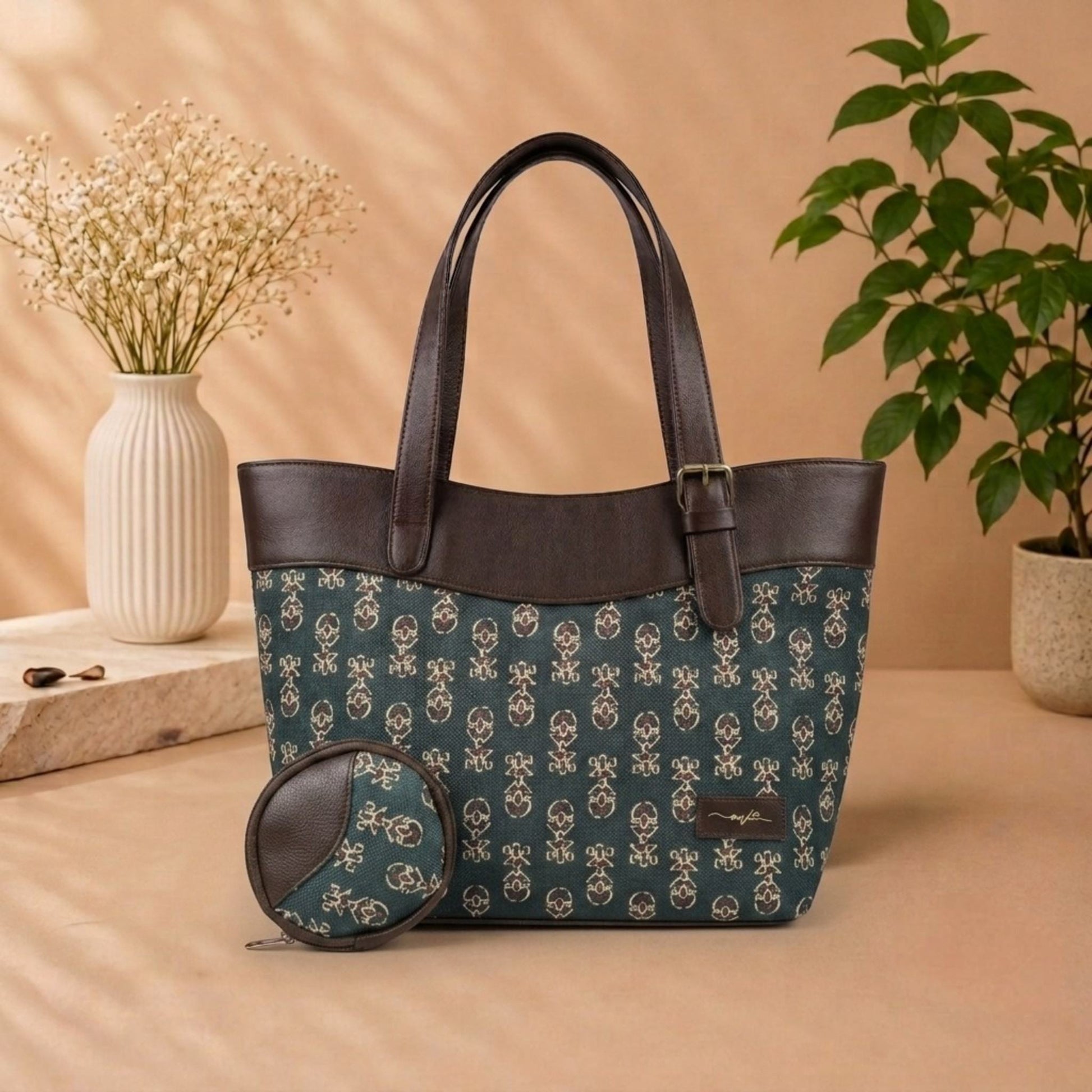 A tote bag with a dark teal and beige floral pattern, dark brown faux-leather trim, and matching zippered coin pouch, styled on a beige surface with a vase of dried flowers and a potted plant in the background.
