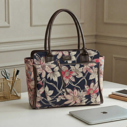 A structured tote bag with a navy blue floral print and dark brown leather handles, styled on a white desk next to a laptop in a home office setting.