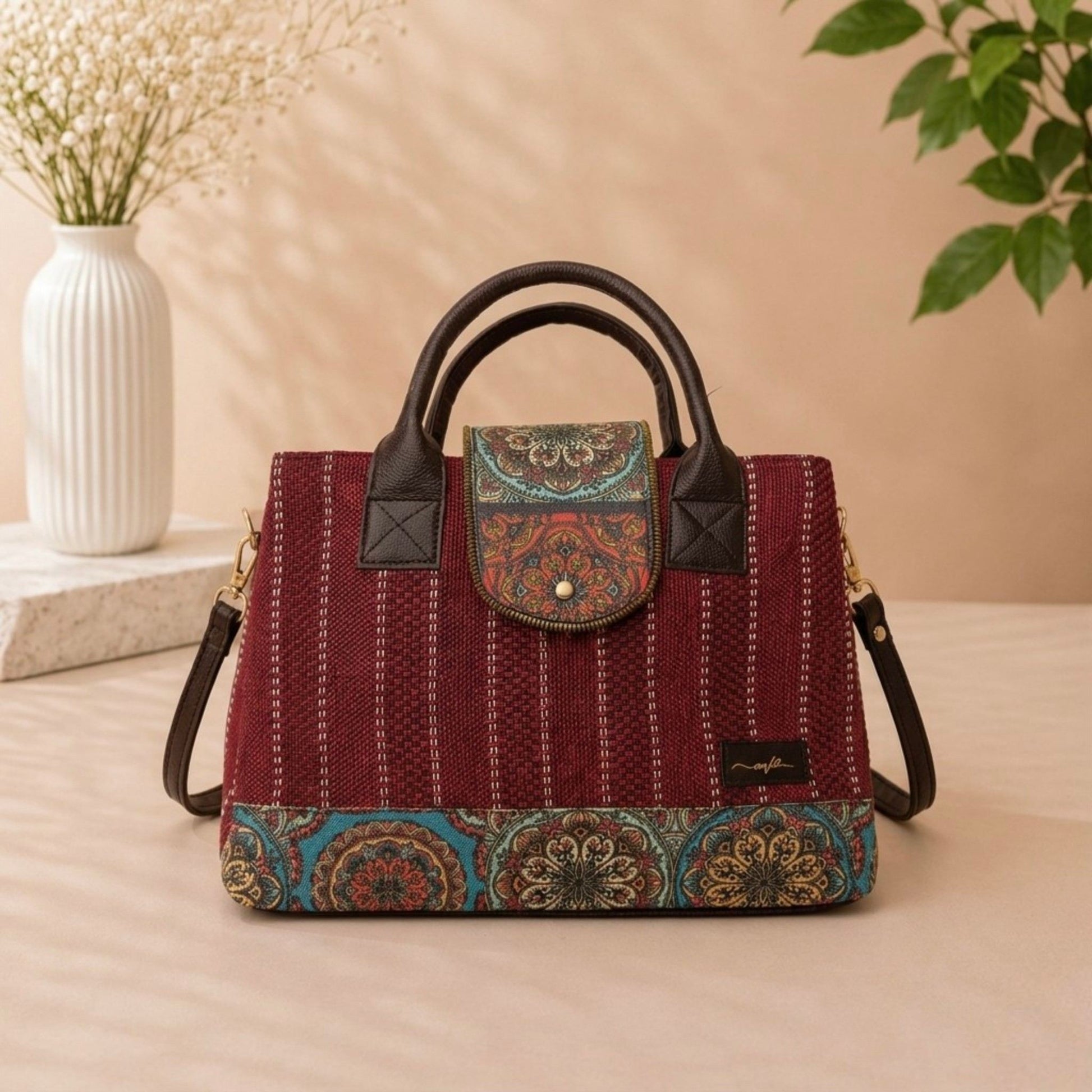 Red patterned handbag with black handles on a beige surface, with a vase and plant in the background.