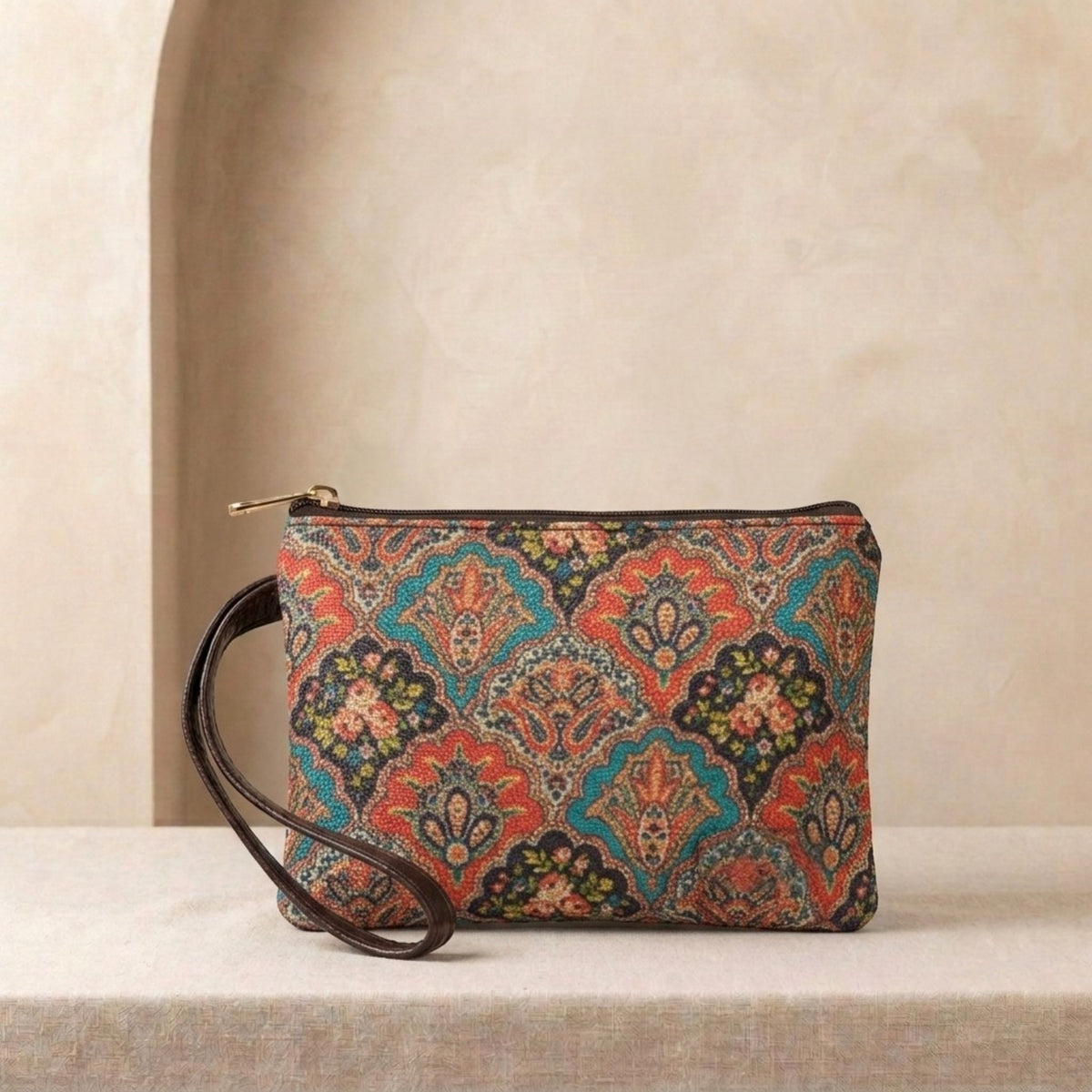 A rectangular clutch bag with a colorful ornate paisley pattern in red, teal, orange, and black, featuring a dark brown leather wristlet strap and a gold zipper, displayed on a textured beige surface against a neutral beige background.
