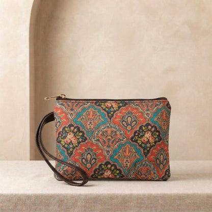 A rectangular clutch bag with a colorful ornate paisley pattern in red, teal, orange, and black, featuring a dark brown leather wristlet strap and a gold zipper, displayed on a textured beige surface against a neutral beige background.