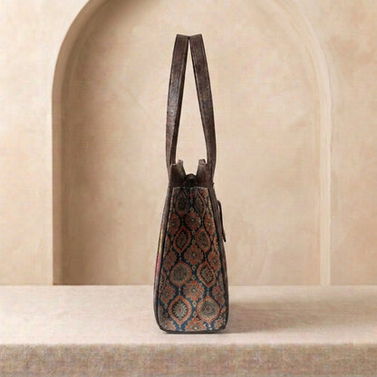 A structured leather shoulder bag with a dark brown patterned body and double shoulder straps, standing upright on a stone ledge against a beige arched background.
