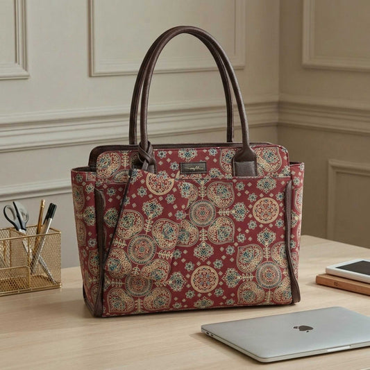 A structured tote bag with a red and beige medallion pattern and dark brown handles, displayed on a light wood desk next to a laptop in a home office setting.