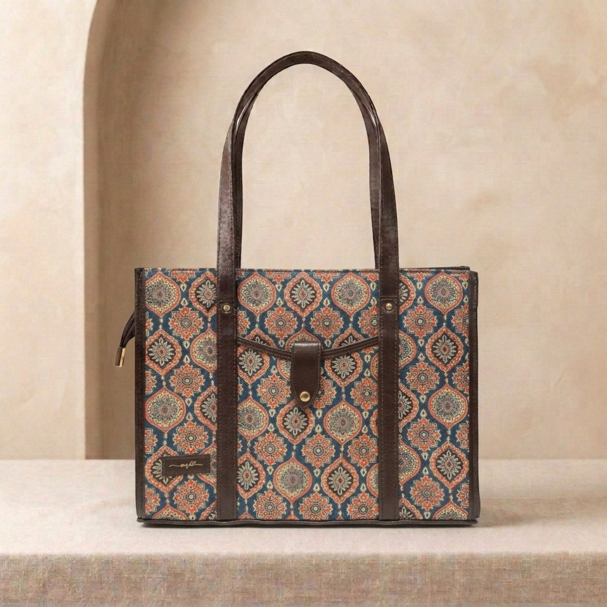 A structured tote bag with a brown leather handle and trim, featuring a detailed geometric medallion pattern in blue, rust orange, and beige on the body. The bag is displayed on a textured beige surface against a matching beige background with a soft arch detail.