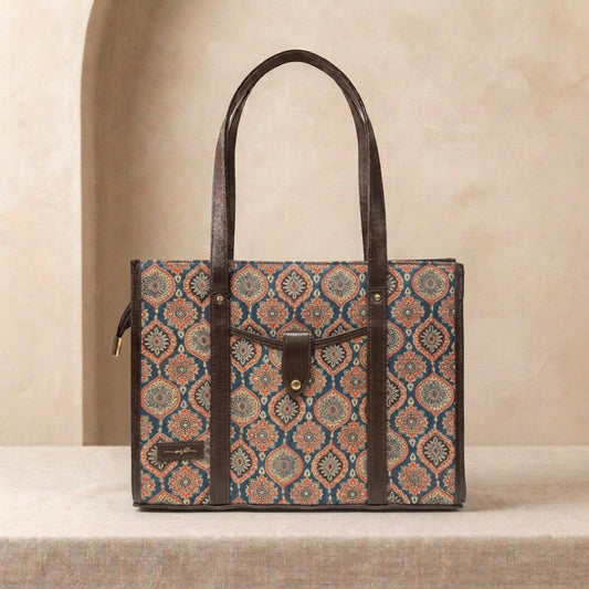 A structured tote bag with a brown leather handle and trim, featuring a detailed geometric medallion pattern in blue, rust orange, and beige on the body. The bag is displayed on a textured beige surface against a matching beige background with a soft arch detail.