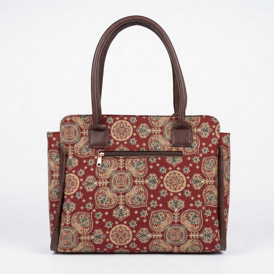 A structured tote handbag with a red background and an intricate beige, blue, and green geometric and floral pattern. It features dark brown faux-leather handles and trim, and a front zippered pocket.