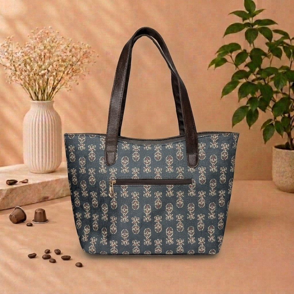 A tote bag with a blue and white monogram pattern and dark brown handles, styled on a beige surface with coffee beans, a vase of flowers, and a potted plant in the background.