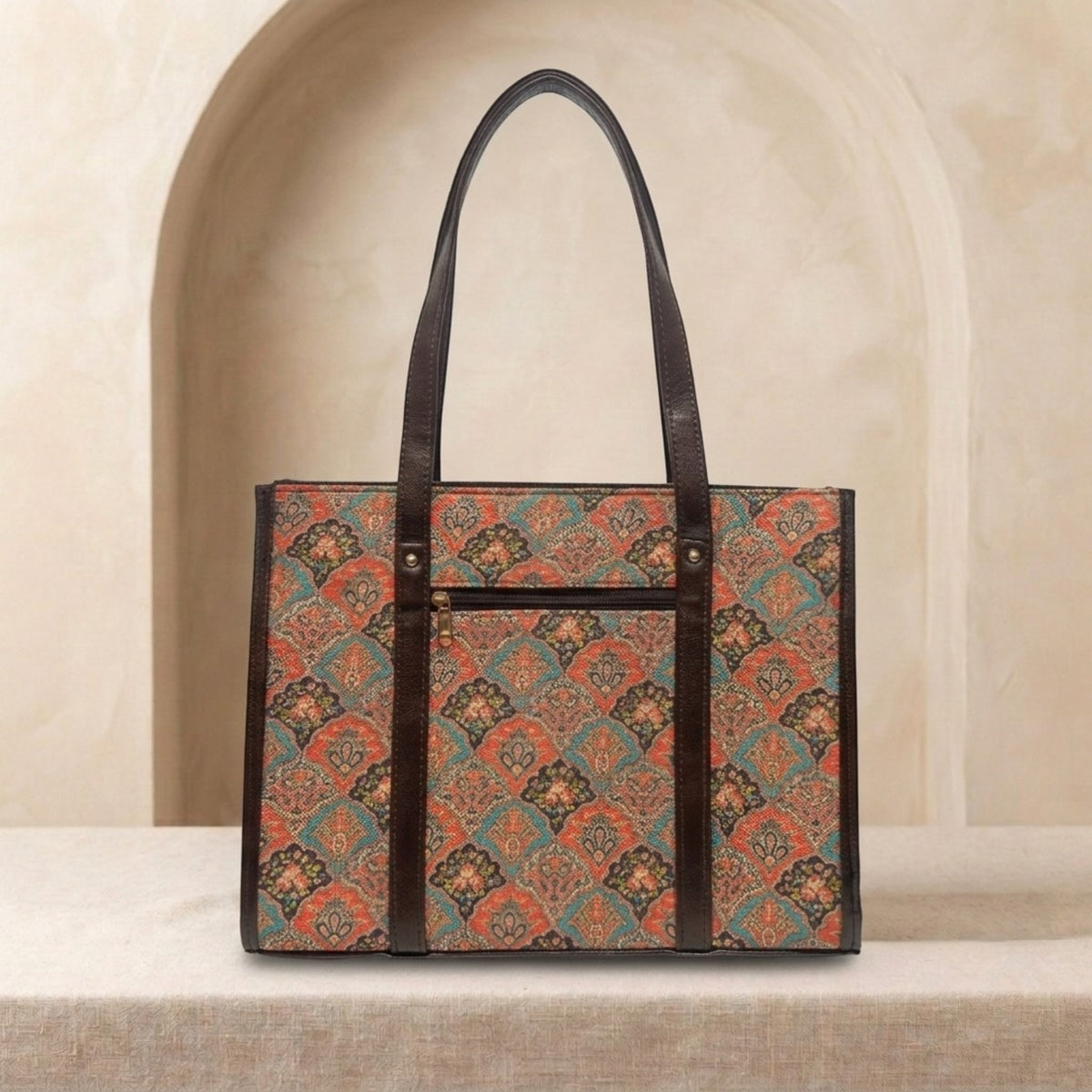 A structured tote bag with a brown leather trim and handles, featuring a vintage-style geometric pattern in rust orange, teal, and brown. The bag is displayed on a textured beige surface in front of a soft beige arched background.