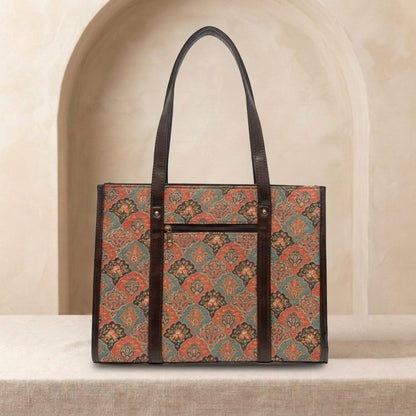 A structured tote bag with a brown leather trim and handles, featuring a vintage-style geometric pattern in rust orange, teal, and brown. The bag is displayed on a textured beige surface in front of a soft beige arched background.