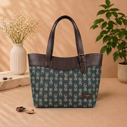 A structured tote bag with dark brown faux leather handles and trim, featuring a teal body covered in a repeating cream floral pattern. The bag is styled on a beige surface with dried flowers in a white vase and a green potted plant in the background.