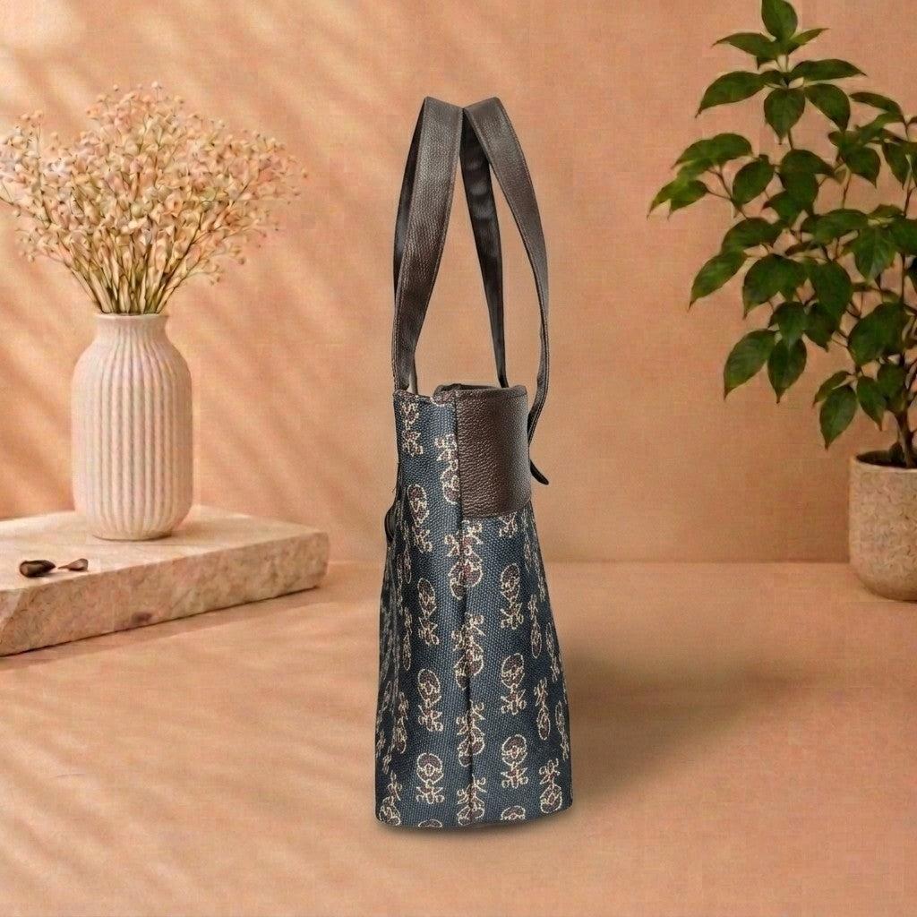 A blue tote bag with a white floral pattern and dark gray straps, standing on a beige surface in a styled room setting with a vase of dried flowers and a potted plant.