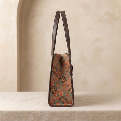 A structured tote bag with dark brown leather handles and trim, featuring a vintage-style geometric pattern in rust orange, teal, and brown, displayed on a neutral beige surface.
