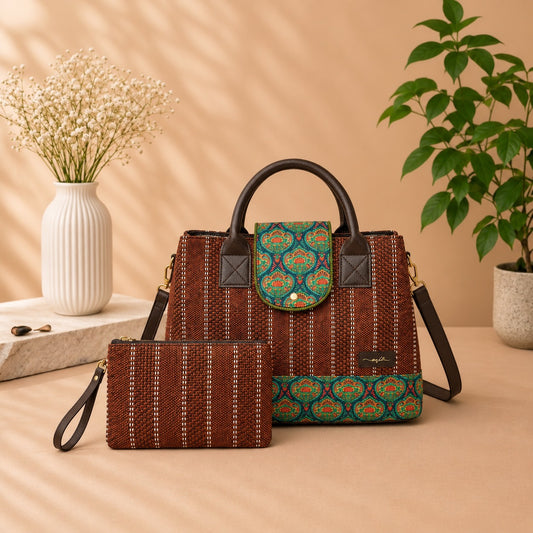 A brown woven handbag with dark brown leather handles and a matching zip pouch placed in front, styled on a beige surface with a vase of dried flowers and a potted plant in the background.